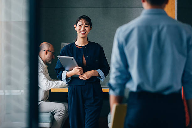 Businesswoman holding a tablet in a professional office environment, engaging with a colleague while another individual works in the background, fostering a collaborative and inclusive workspace.